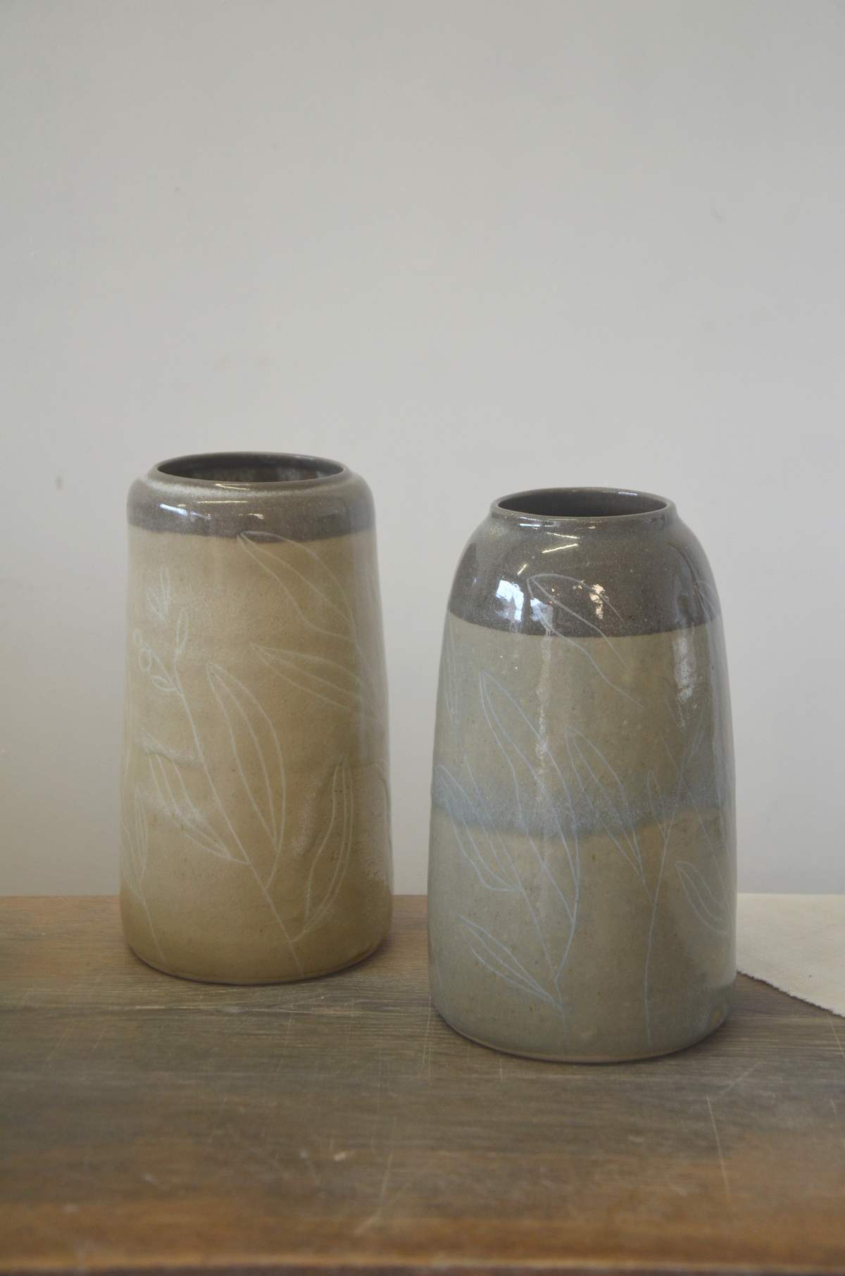 Deux vases à col gris, décor de feuillages clairs, posés sur une table en bois.