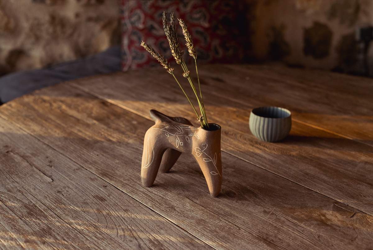 Pièce brune à quatre pieds portant des graminées, un bol clair à côté, sur une table en bois devant un mur de pierre.