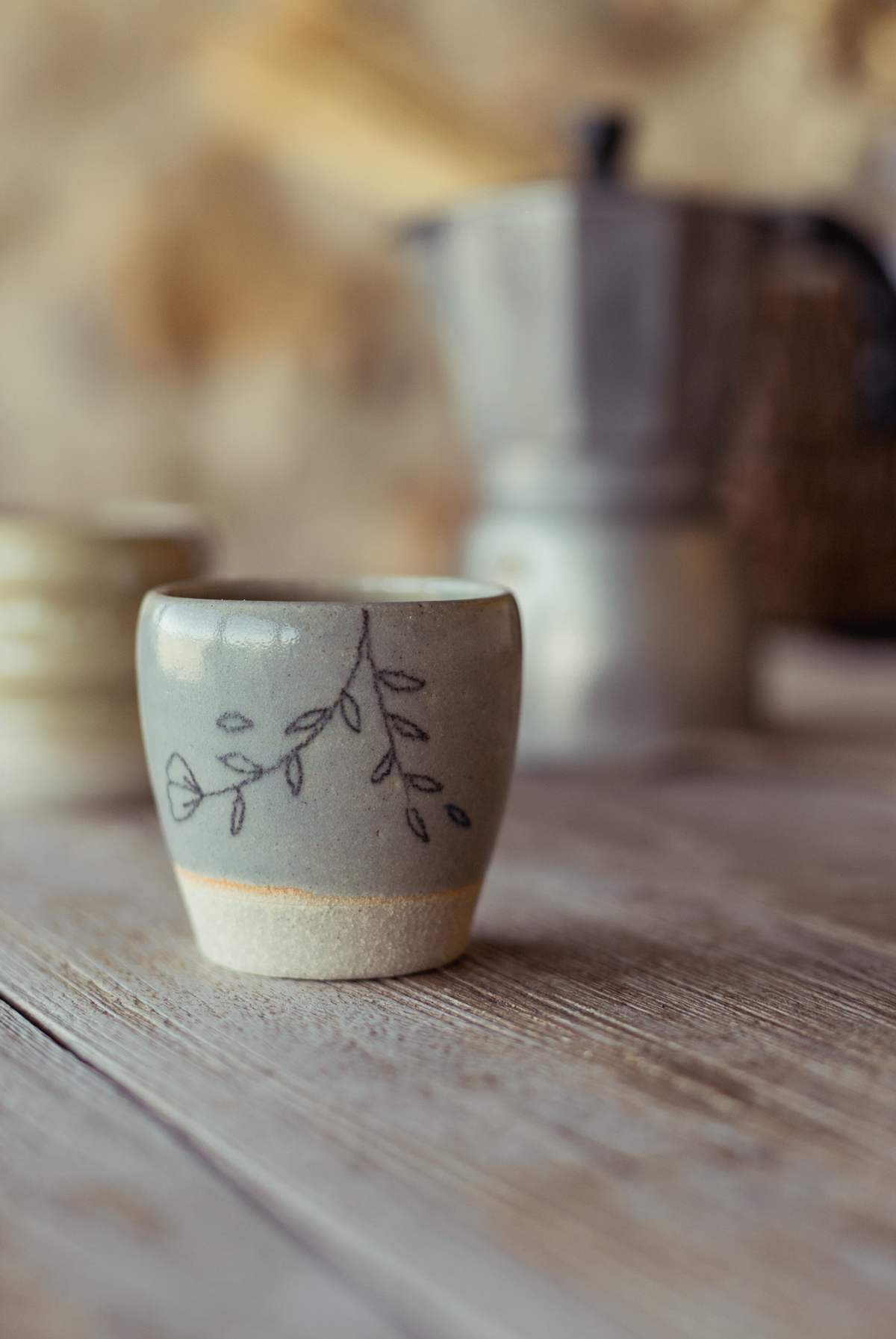 Tasse claire décorée de brins de feuillage, cafetière floue en arrière-plan.